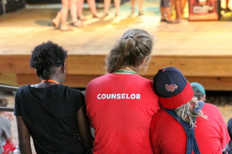 Three people sit in a row facing away from the camera. The middle person is wearing a shirt that says COUNSELOR on the back, and the person on the right is leaning her head on the counselor's shoulder.