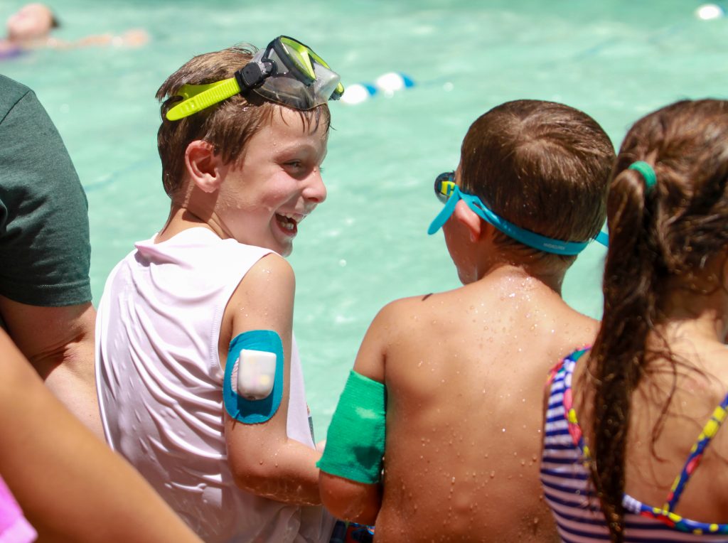Two boys sit and laugh at a pool. The boy on the left has an Omnipod on his upper arm.