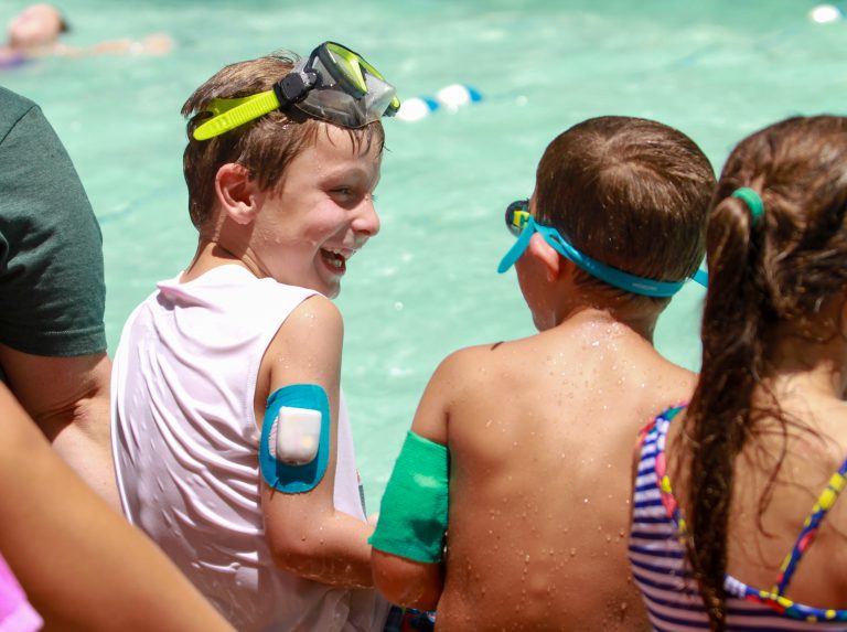 Two boys sit and laugh at a pool. The boy on the left has an Omnipod on his upper arm.